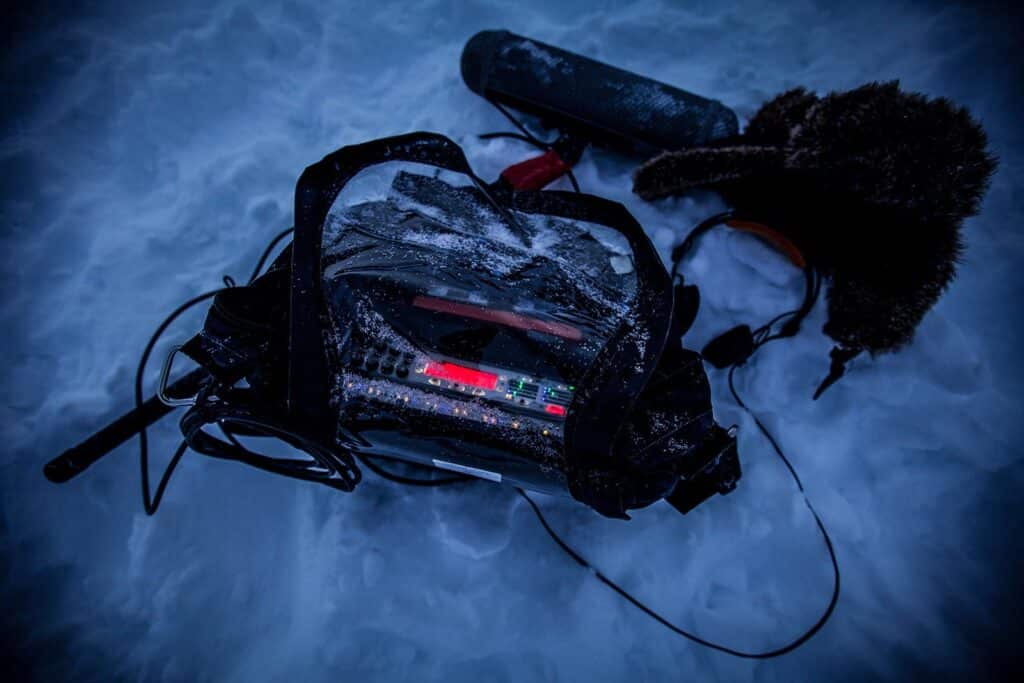 A portable audio recorder for sound effects libraries sits on snowy ground with cables, headphones, and a furry hat.