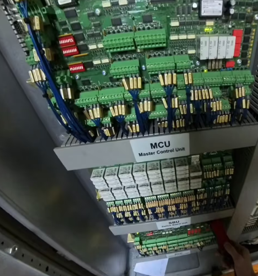 Electrical control panel with "MCU Master Control Unit" and "SBU" labels, green circuit boards, wiring, relays; hand adjusts part below.