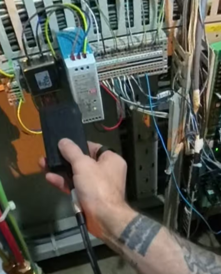 A tattooed person uses a handheld device connected by cable to a control panel with wires, performing maintenance or diagnostics.