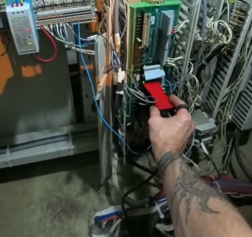 A tattooed person’s hand attaches a red device to an open electrical control panel with exposed wires, circuit boards, and cables.
