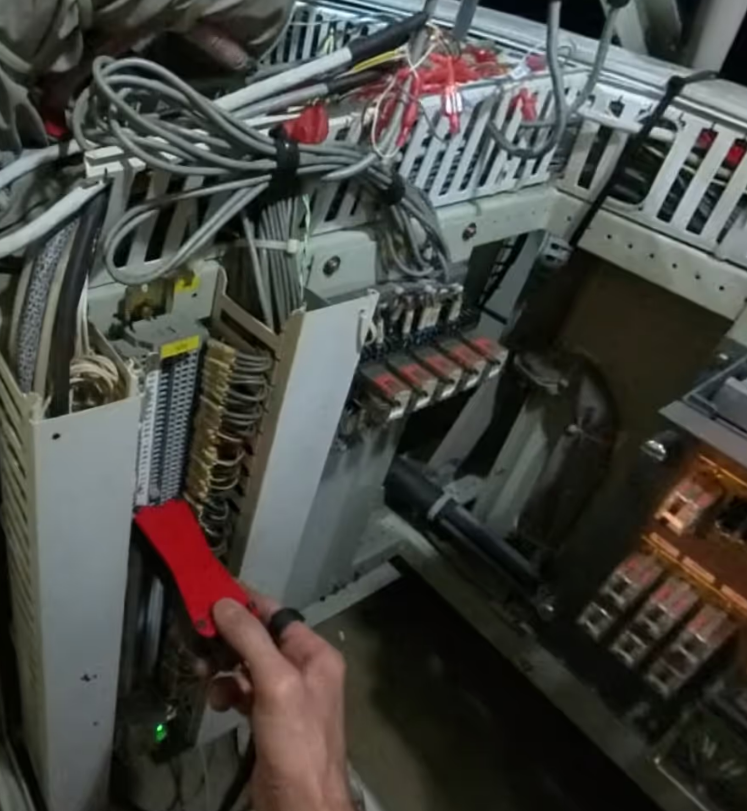 A person uses a red handheld tool to work on wiring inside an open electrical control panel containing cables, relays, and connectors.