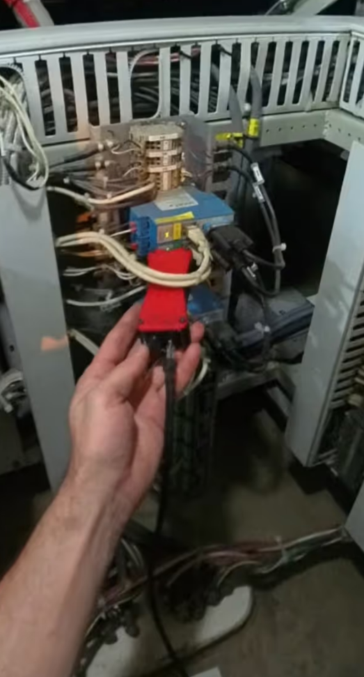 Hand holding a red connector with cable near an open electrical control panel filled with wires and electronic components.