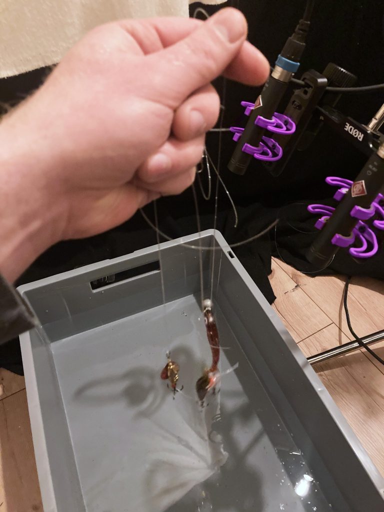 A hand holds fishing lures above a gray water-filled tub, with microphones and pop filters in the purple-lit background.