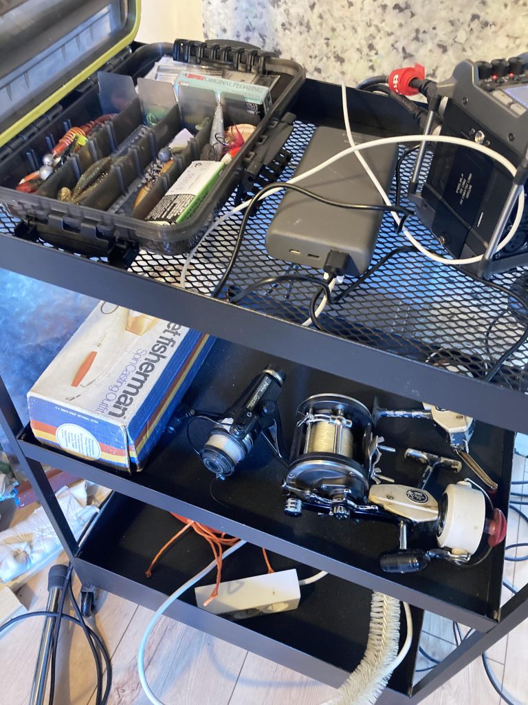 Black metal cart with shelves holds fishing reels, tackle box, power strip, cables, tissue box, and tools in a well-lit room.