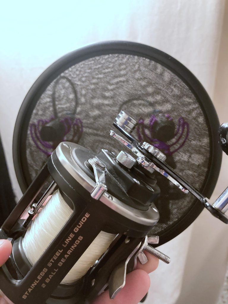 A fishing reel with white line held in front of a round pop filter featuring a purple spider web design on a light background.