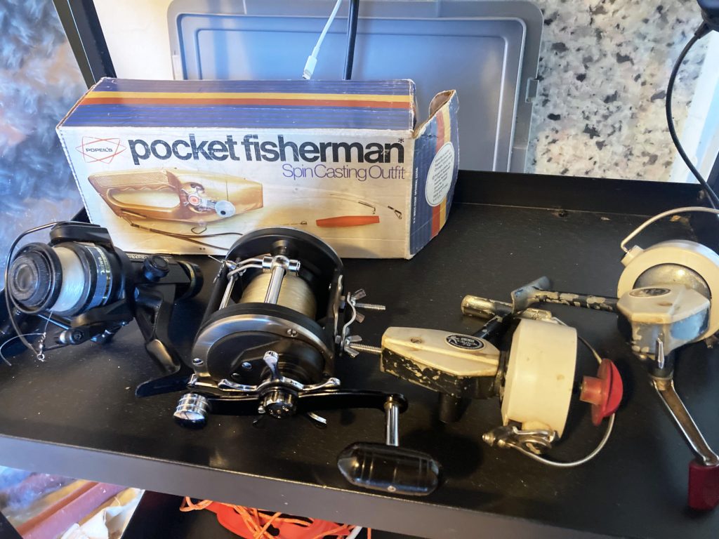 Four vintage fishing reels on a black shelf, with a boxed "Pocket Fisherman Spin Casting Outfit" behind them. Gray container and granite-patterned background.