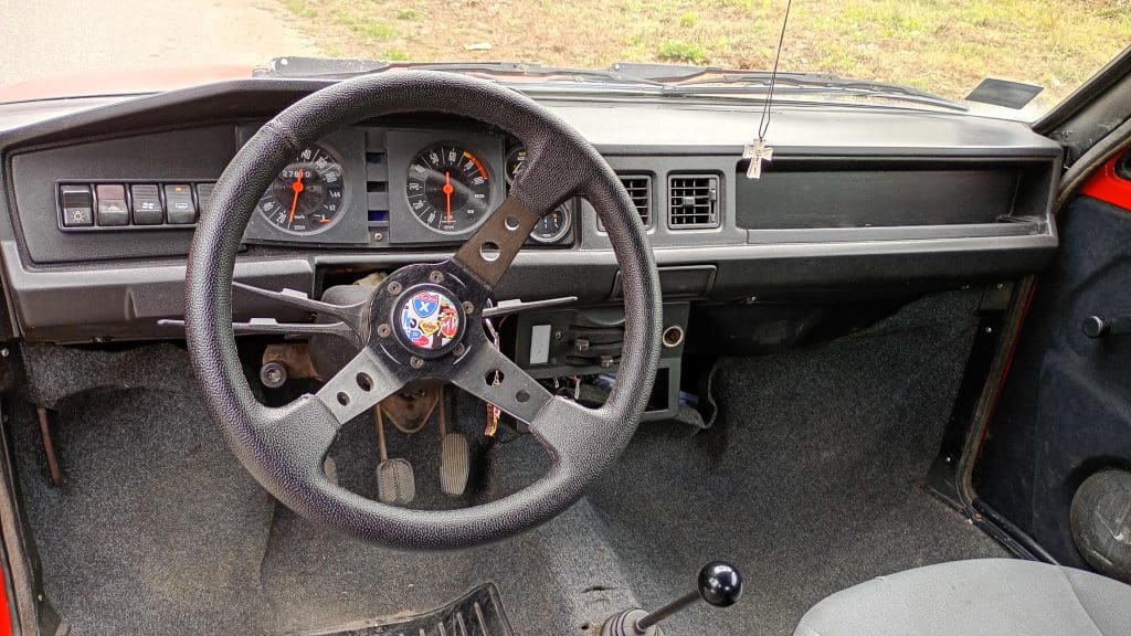 Vintage car interior featuring dashboard, steering wheel with emblem, instrument gauges, ventilation controls, gear stick, and pedals.