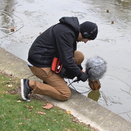 A person in a black hoodie and beanie kneels by a pond, using a furry microphone to record sounds. Headphones and red bag visible.