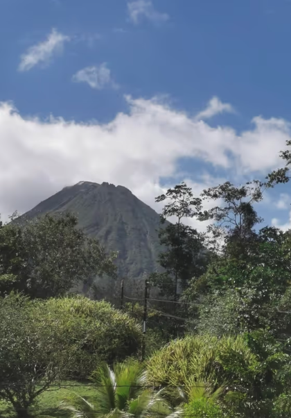 Lush green landscape with trees and shrubs in the foreground, tall cone-shaped volcano in background beneath partly cloudy blue sky.
