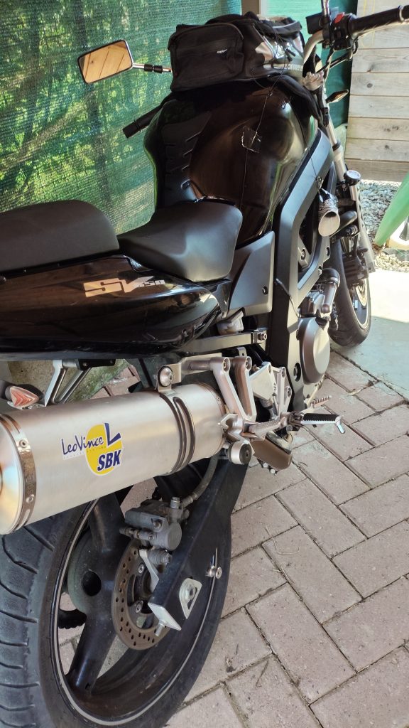 A black motorcycle equipped with a LeoVince SBK exhaust is parked on a brick surface beside a green mesh fence and several wooden planks.