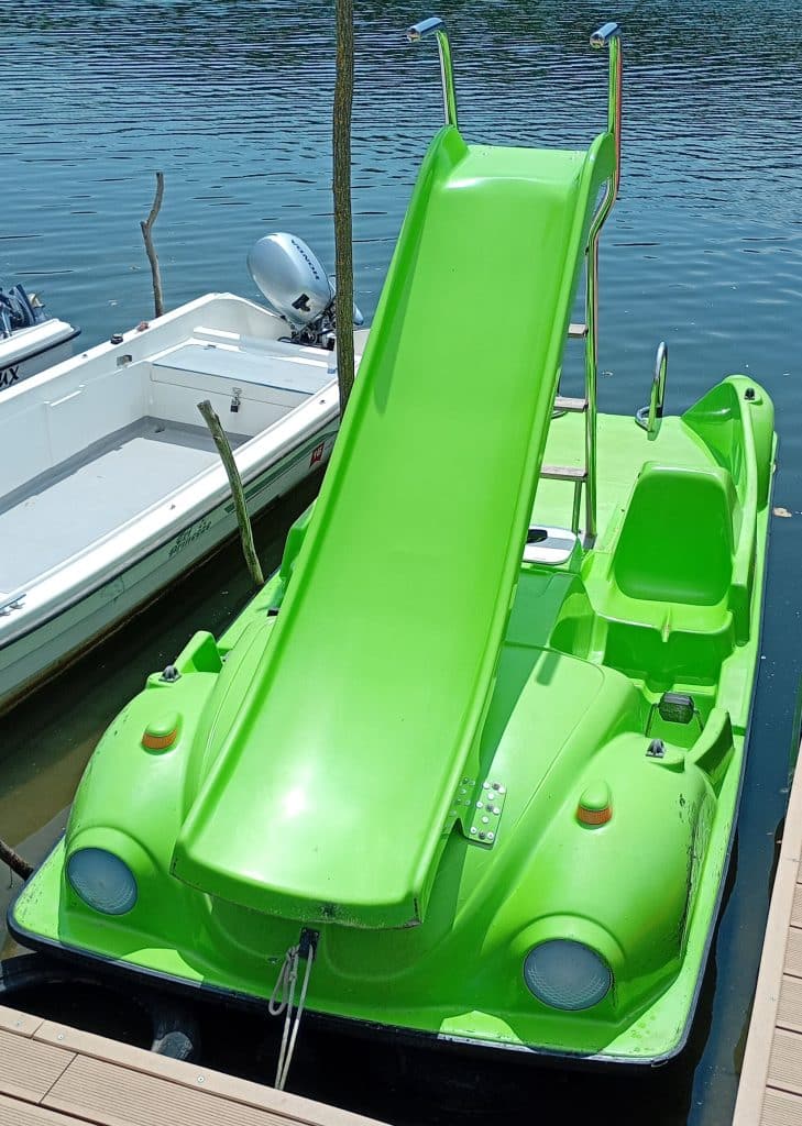 A bright green pedal boat with a built-in slide is docked on the water beside a white motorboat. The pedal boat features headlights and a unique design that resembles an old car.