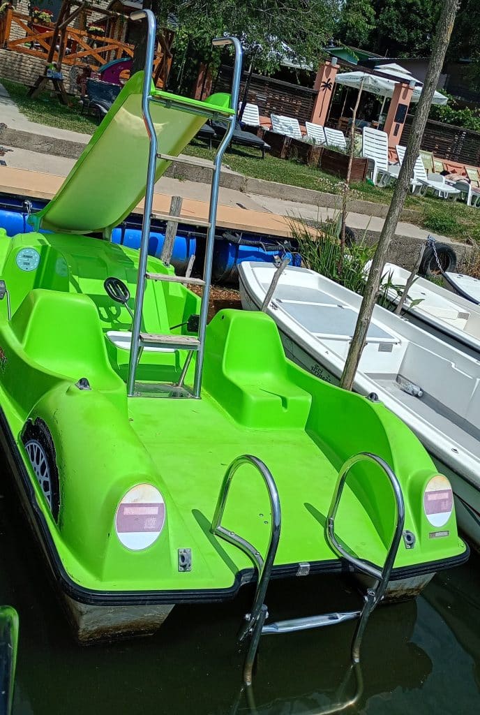 A bright green pedal boat with a slide and metal ladder is docked by the shore next to a white boat. Lounge chairs and a small building appear in the background.