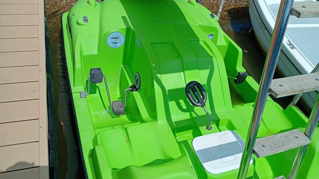 A bright green pedal boat is docked beside a wooden pier, featuring two black pedals, a steering wheel, and a white cooler resting on the seat. On the right side, part of a white boat and a metal ladder are visible.