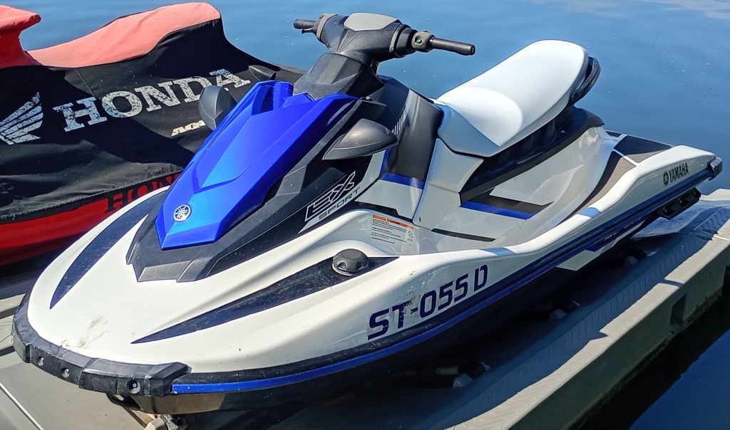 A blue and white Yamaha jet ski rests on a floating platform beside a covered Honda jet ski, with water in the background.