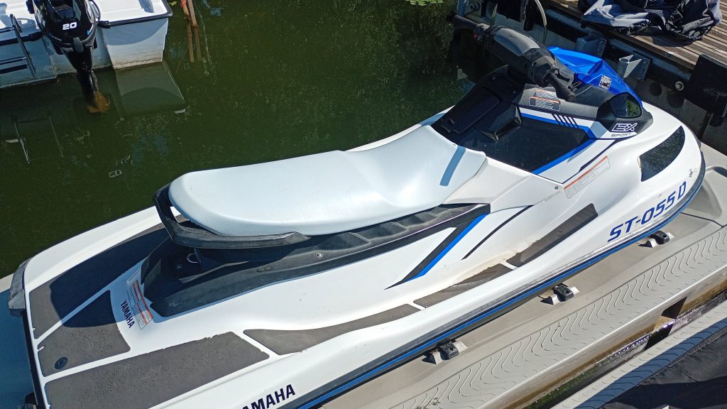 A white and blue Yamaha jet ski with black handlebars is docked next to a wooden pier on calm water, with another boat partially visible nearby.