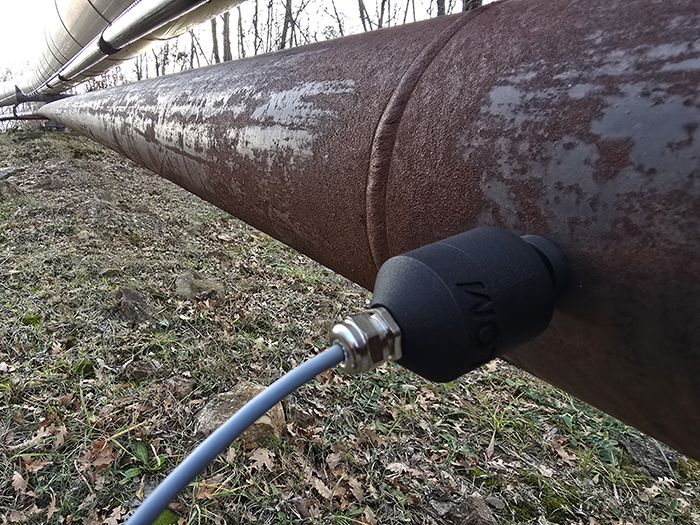 Close-up of a large outdoor industrial pipeline featuring a black sensor device attached to the pipe with a cable extending from it. Grass and leafless trees are in the background.