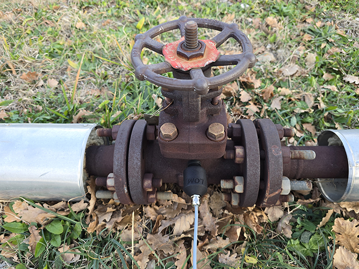 A rusted metal valve with a handwheel connects two large pipes, situated on grassy ground with dry leaves scattered around.