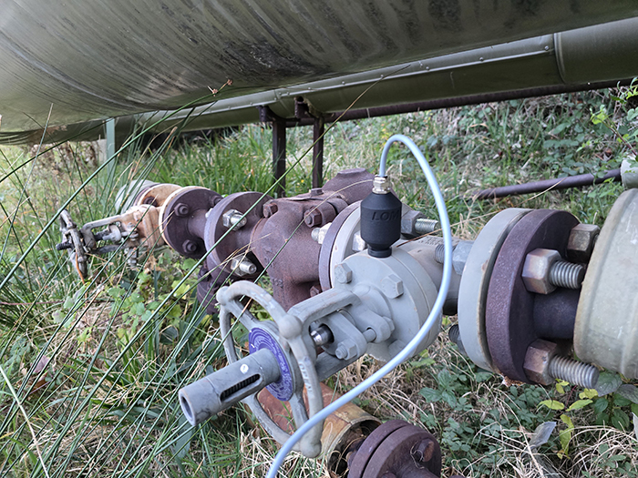 Close-up view of a cylindrical metal pipeline featuring several valves and connectors, running through grassy terrain. The pipe is supported by metal braces, with wiring visibly attached to some of the valves.