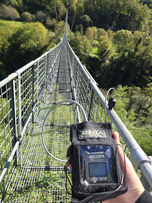 A person holds a Zoom audio recorder on a metal suspension bridge, with green trees in the background and a clear blue sky above.