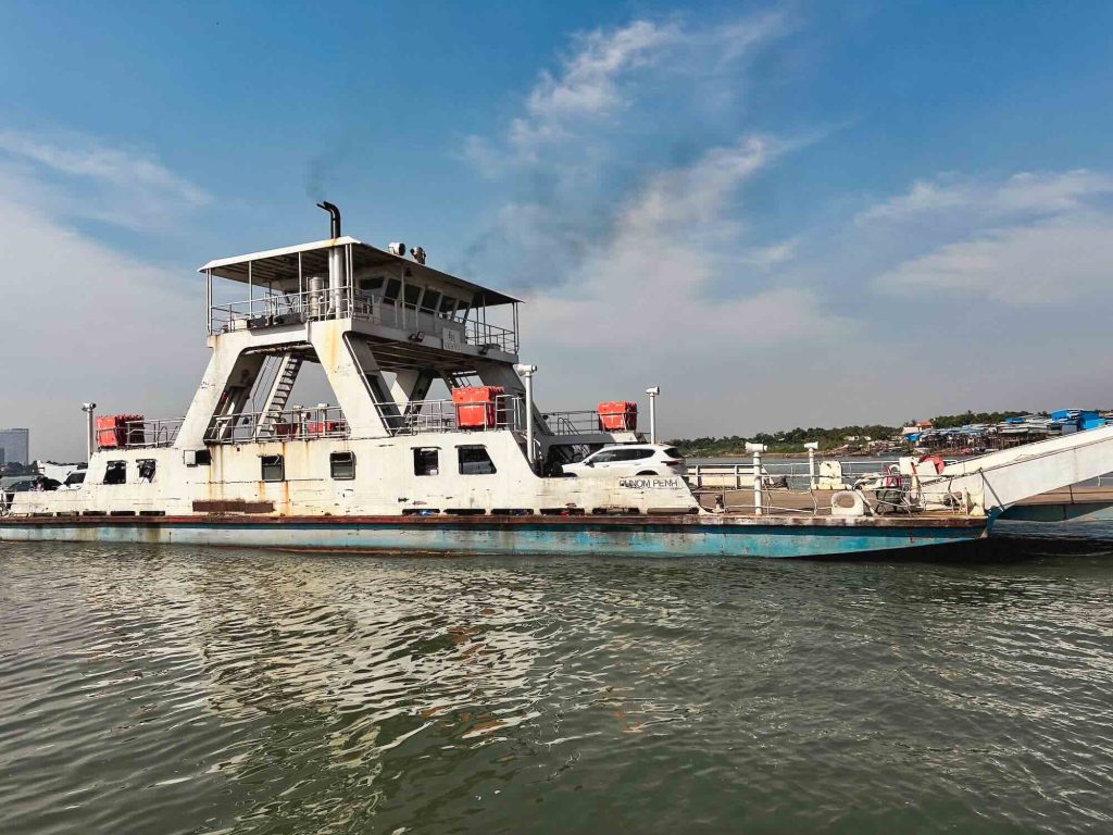 A white ferry equipped with orange lifebuoys is docked beside a ramp on calm water. In the background, a partly cloudy blue sky stretches over a shoreline lined with buildings and greenery.