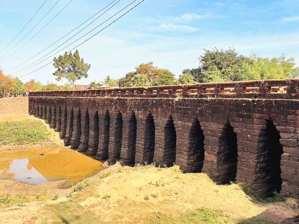 A historic stone bridge with multiple arches spans a dry, shallow riverbed. Grass, trees, and distant houses surround the area, while power lines cross overhead against a blue sky.