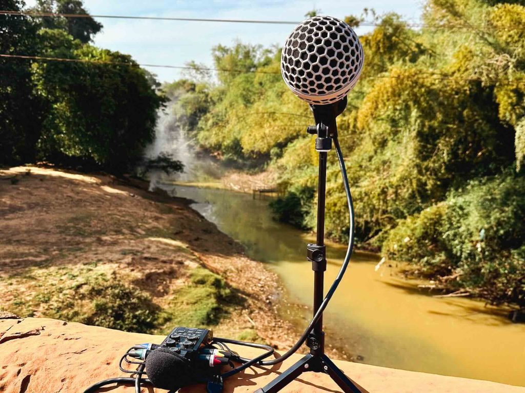 A microphone on a stand with an audio recorder is positioned outdoors beside a muddy river, surrounded by trees and foliage under a clear sky.