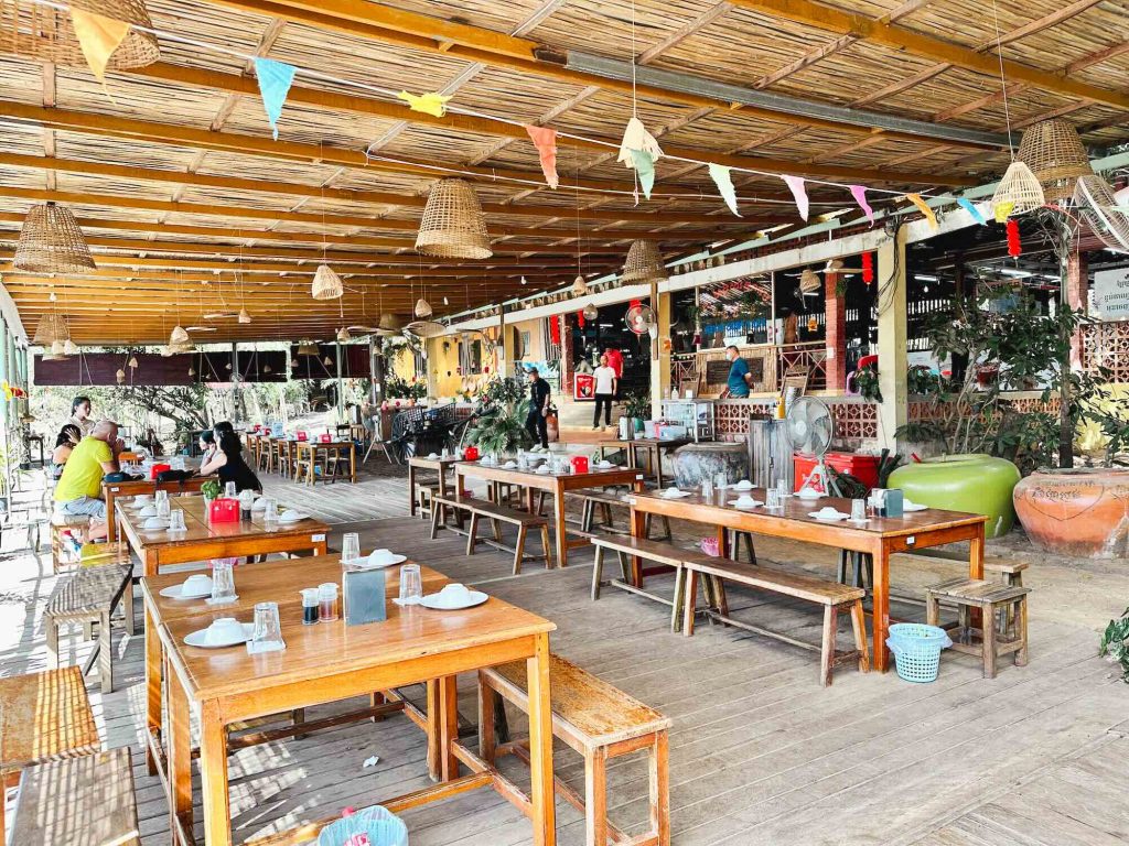 Rustic open-air restaurant featuring wooden tables and benches, hanging lights, colorful flags, and several diners seated. The setting is bright and airy, with plants and various decorations surrounding the space.