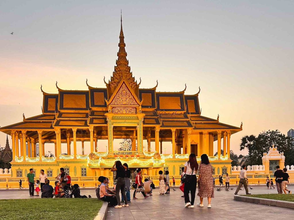 People gather in front of a golden traditional pavilion with ornate roof details at sunset, under a clear sky. Some individuals walk, sit, or take photos, interacting with the setting in a calm cultural environment.