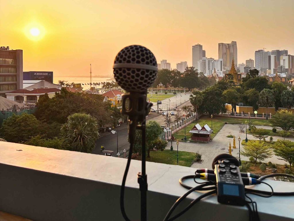 A microphone and audio recorder are set up on a balcony overlooking a cityscape at sunset, with tall buildings, green areas, and a park in the background.