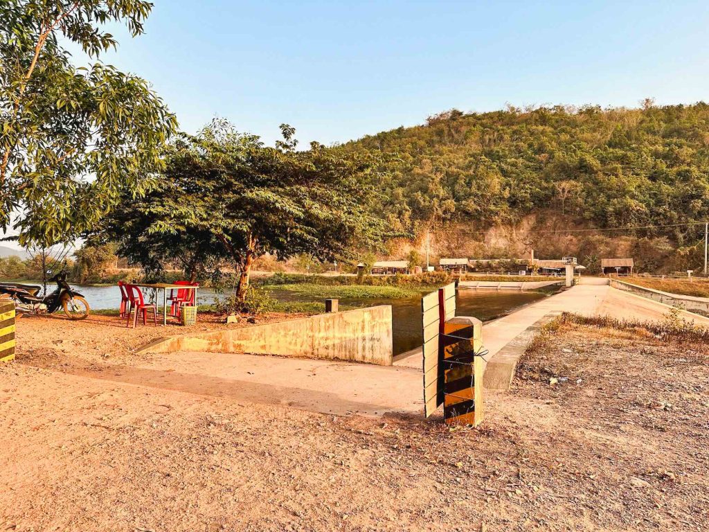 Rural riverside setting featuring a tree, red chairs and a table near the water, a motorcycle parked nearby, a concrete bridge crossing the river, and a green hill in the background under a clear sky.