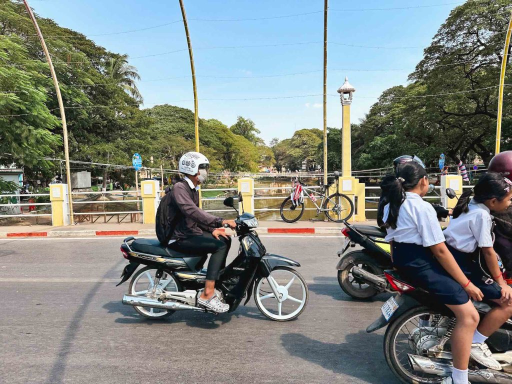 A helmeted person rides a scooter, passing a group of school children sharing another scooter as they cross a bridge surrounded by trees and bordered by a canal.