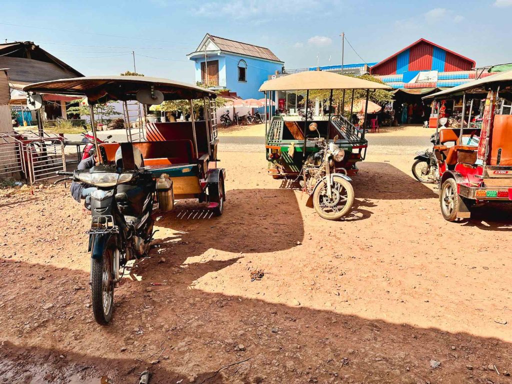 Tuk-tuks and motorbikes parked on a dirt lot beneath bright sunlight, with colorful houses and buildings in the background.