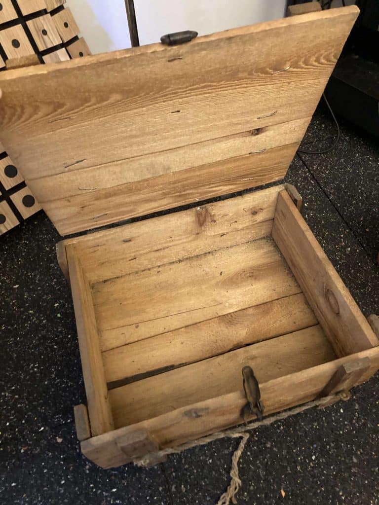 An open, empty wooden box with a hinged lid rests on a speckled black floor. The box shows visible wood grain and has a latch in front, giving it a rustic, handmade appearance.