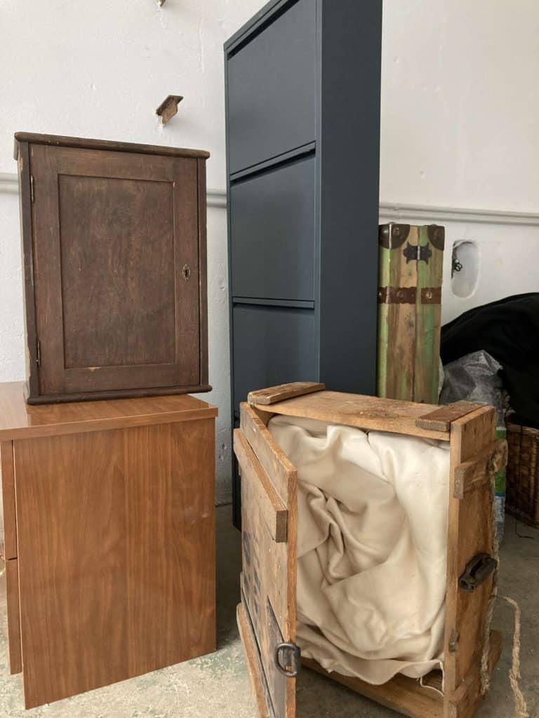 Wooden cabinets and boxes, including an open crate lined with fabric, are arranged on a floor against a white wall.