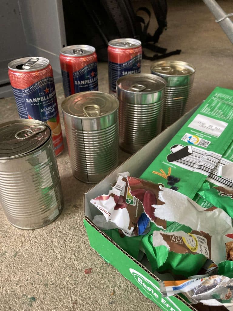 Five empty cans, including two Sanpellegrino cans, and a green recycling bin containing torn wrappers rest on a concrete floor.