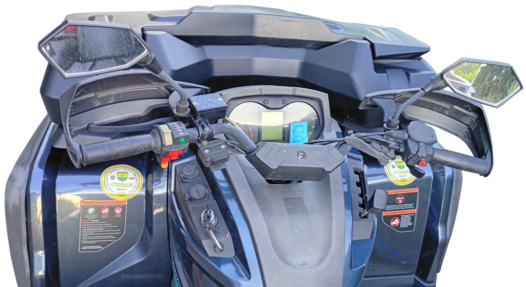 Close-up of an ATV dashboard and handlebars from the rider’s perspective, displaying mirrors, controls, gauges, warning stickers, and various buttons.