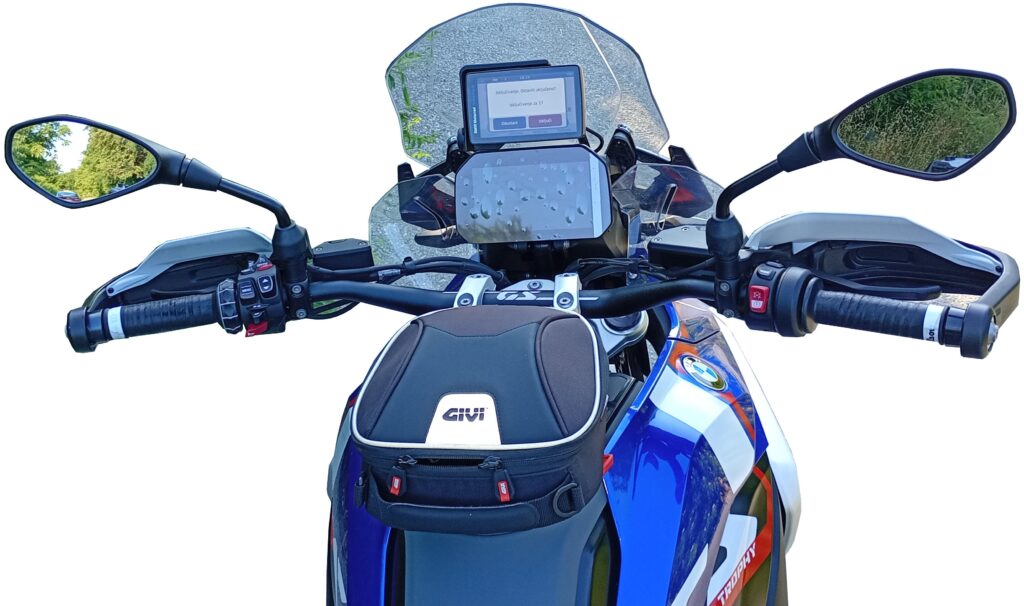View from the rider’s seat of a blue, red, and white motorcycle, showing handlebars, mirrors, a mounted GPS device, and a black Givi tank bag. The background is white.
