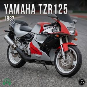 A 1987 Yamaha TZR125 motorcycle in red, black, and silver is parked on a racetrack. The bike has a sporty fairing and “Racing” lettering on the side.