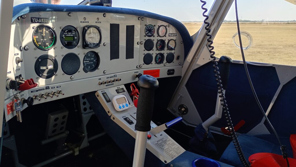 Cockpit view from a small aircraft showing the instrument panel with analog dials, a central control stick, and a view of an open field through the front windshield.