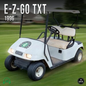A white electric 1996 E-Z-Go TXT golf buggy with headlights on drives across a grass course.